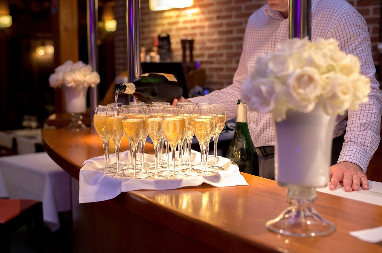 Un serveur verse du champagne dans des flûtes alignées sur un comptoir en bois, avec des fleurs blanches en premier plan.