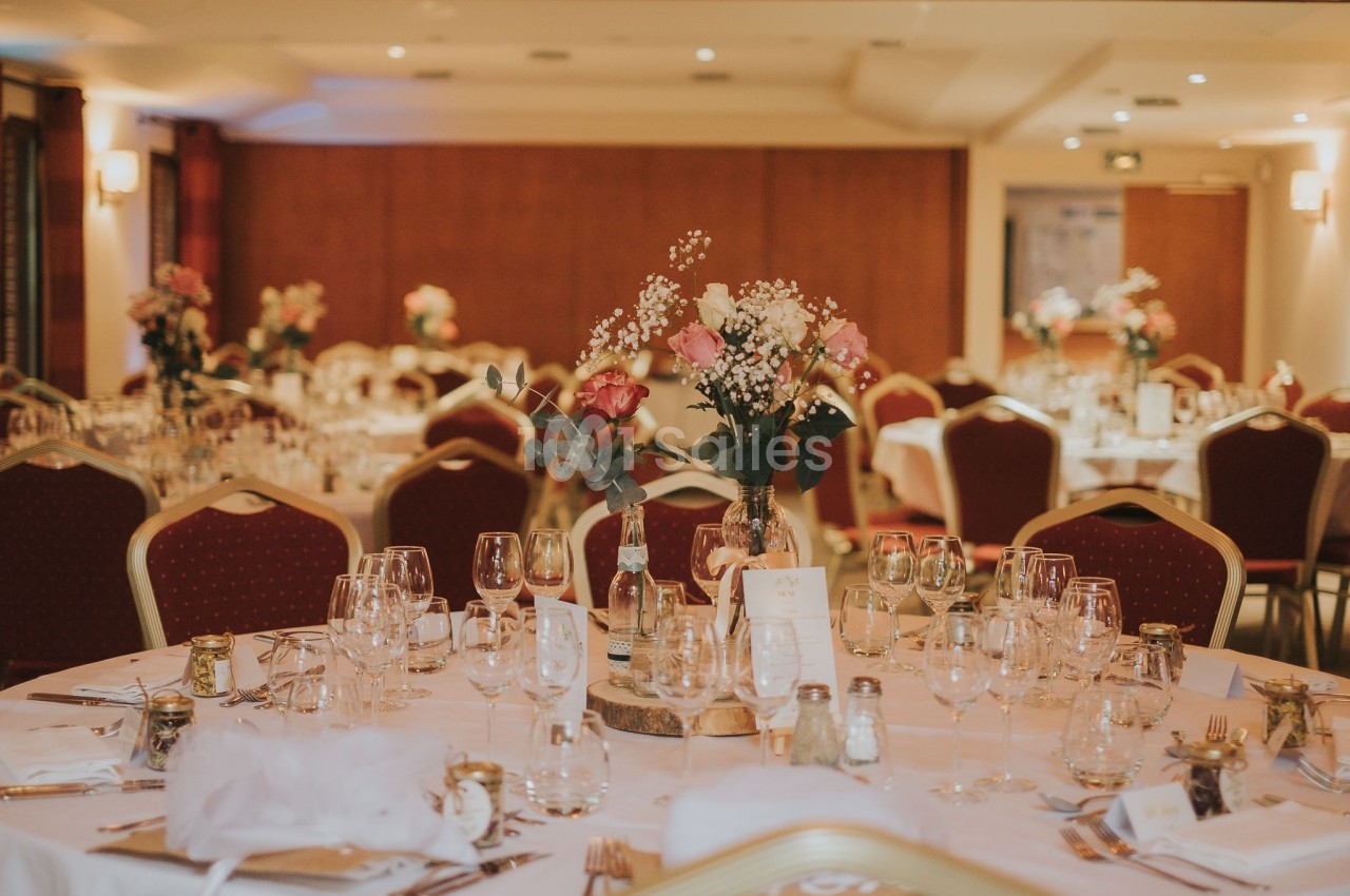 Salle de réception décorée avec des tables rondes, nappes blanches, bouquets de fleurs et verres disposés.