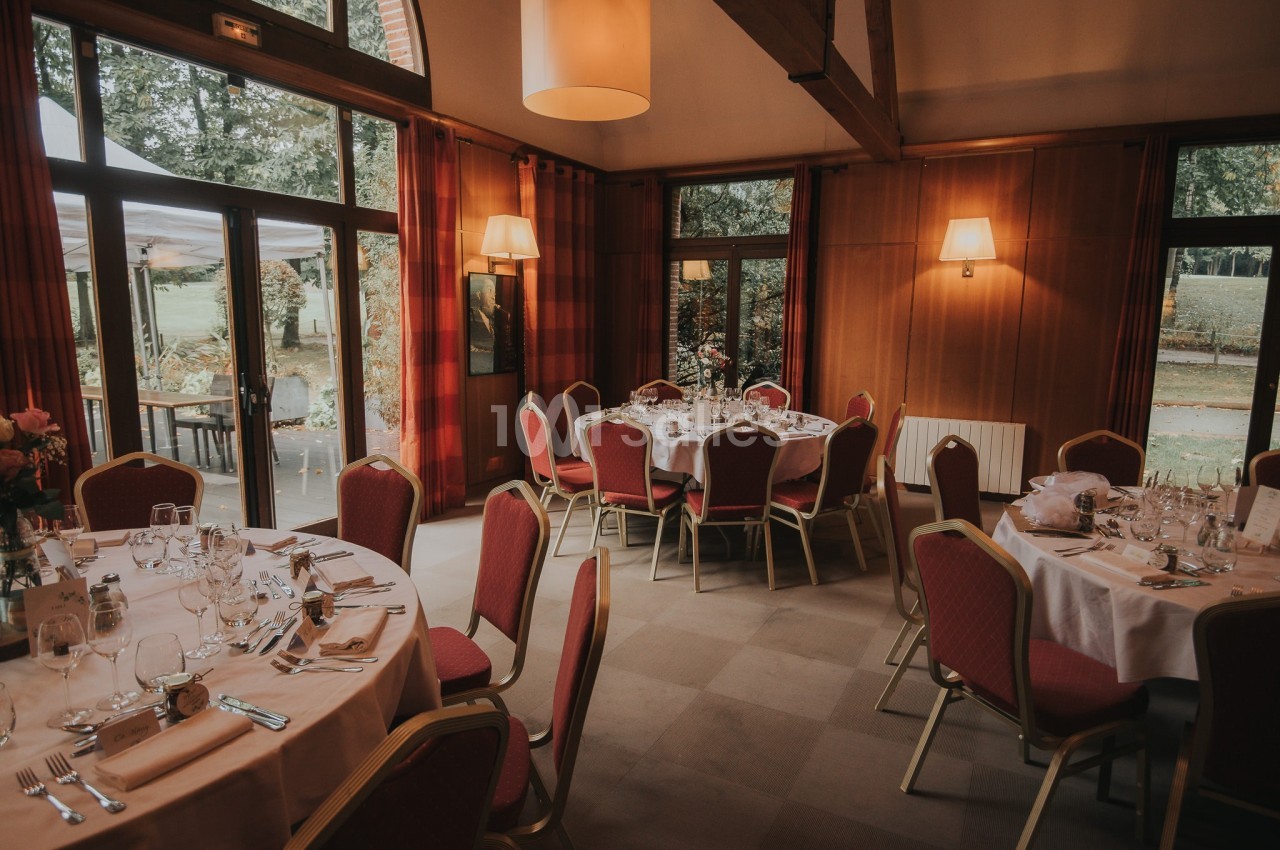 Salle de réception avec tables rondes dressées, chaises rouges et grandes fenêtres donnant sur un jardin.
