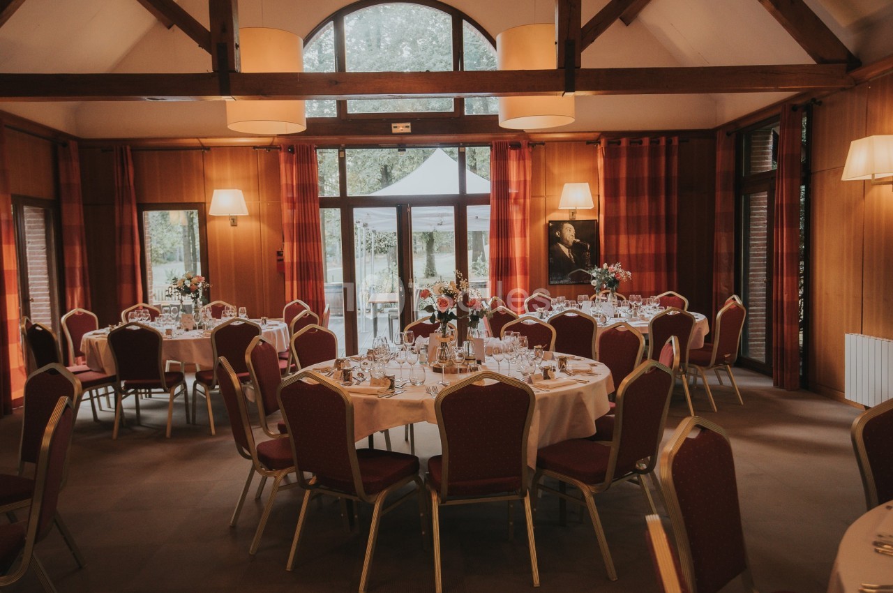 Salle de réception avec tables rondes dressées, chaises rouges, poutres apparentes et lumière naturelle.