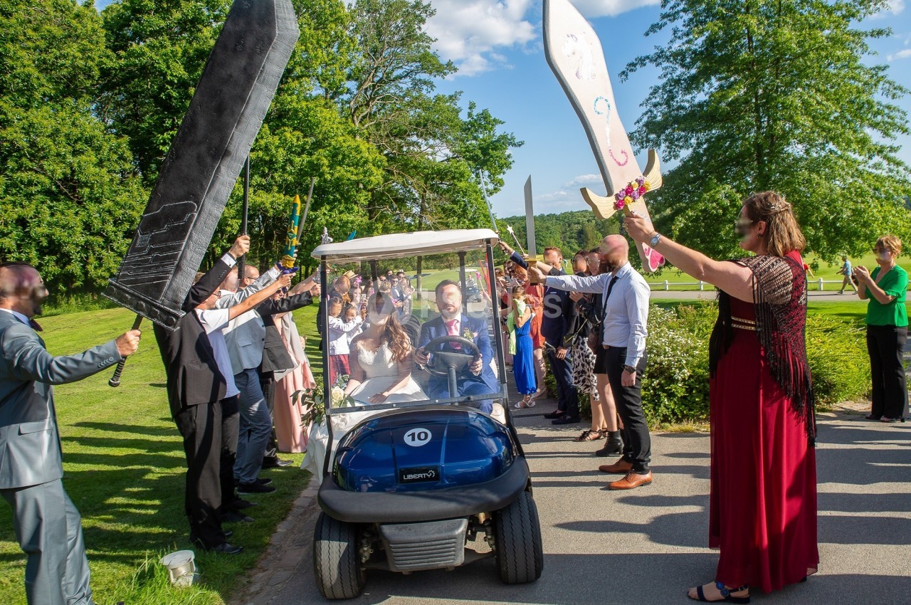 Un homme dans une voiturette de golf passe sous une haie d'honneur formée par des invités brandissant des épées géantes.