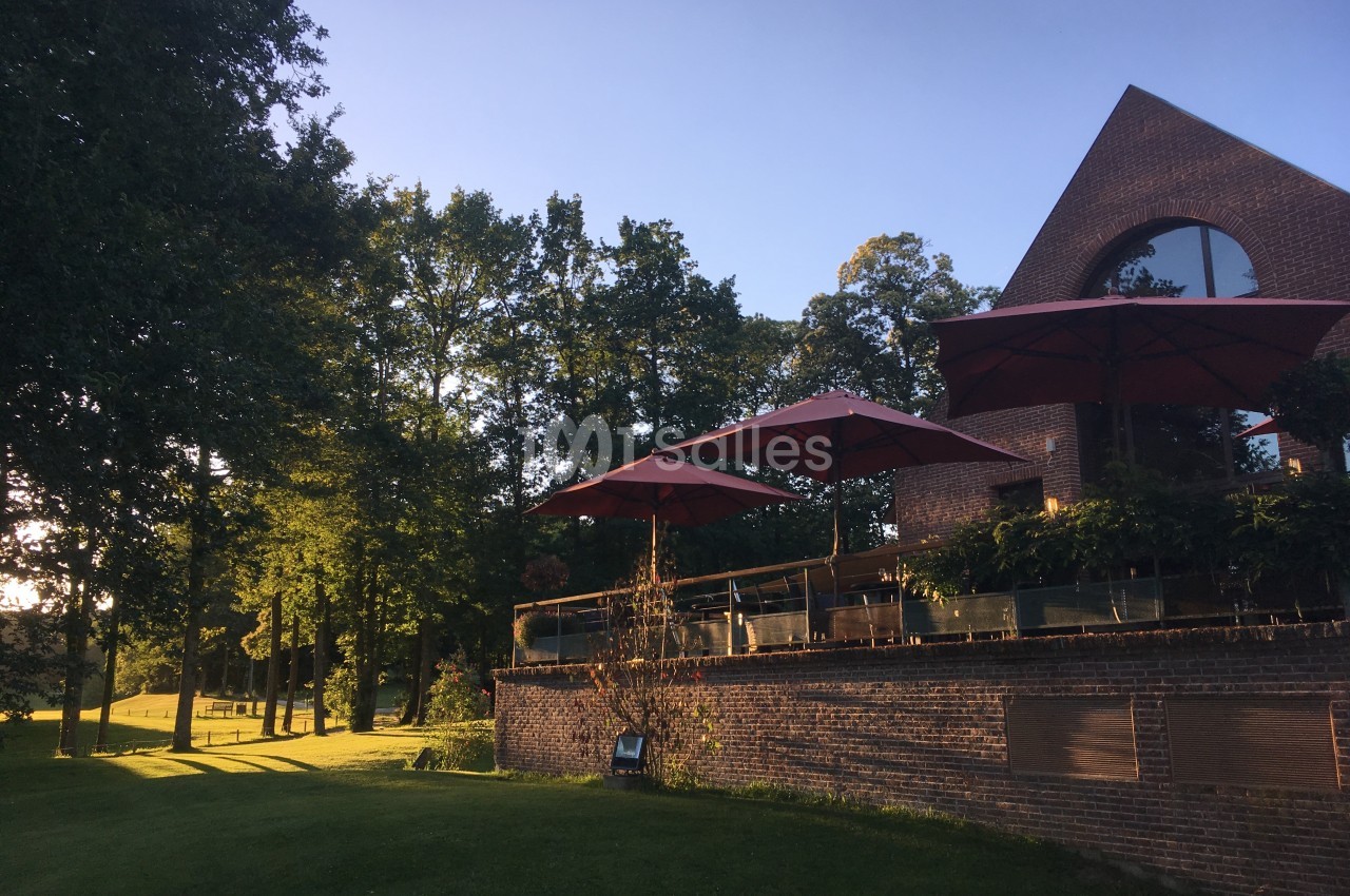 Terrasse en briques avec parasols rouges, entourée d'arbres et d'une pelouse, éclairée par une lumière de fin de journée.
