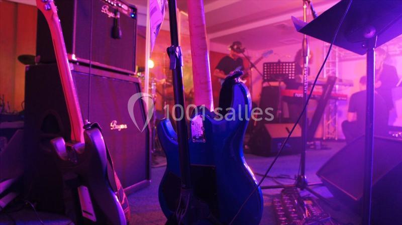Guitare bleue posée sur scène devant des amplificateurs, avec des musiciens jouant en arrière-plan dans une ambiance…