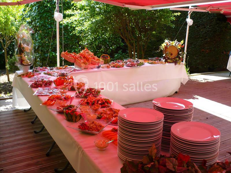 Buffet en extérieur avec plats variés sur des tables blanches et piles d'assiettes sous un auvent rouge.