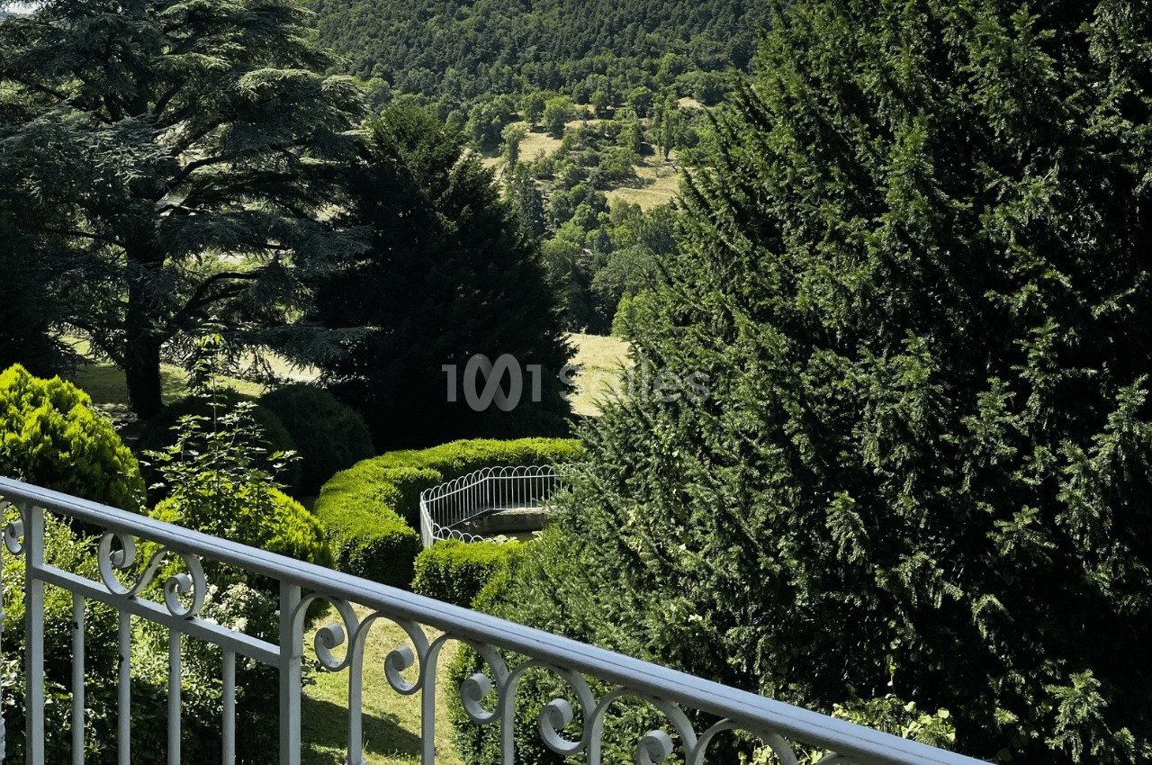 Vue d'un jardin verdoyant avec des arbres, une balustrade blanche au premier plan et des collines en arrière-plan.