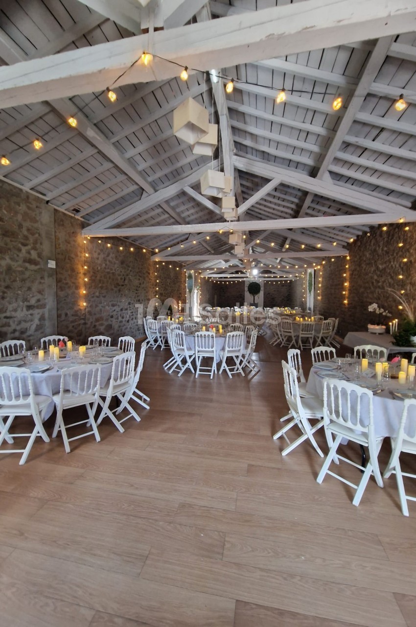 Salle de réception décorée avec des tables rondes, nappes blanches, chaises blanches et guirlandes lumineuses suspendues.