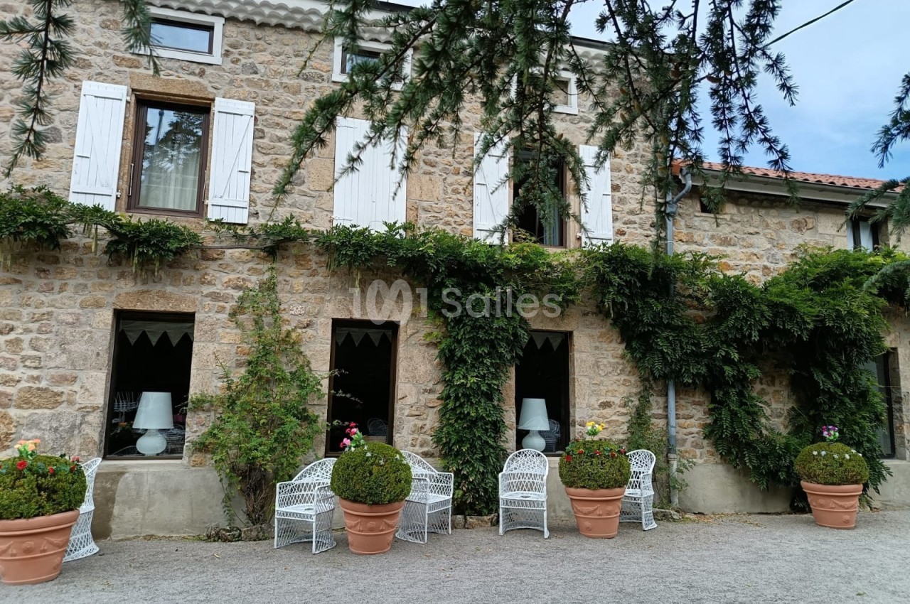 Façade en pierre d'une maison avec volets blancs, plantes grimpantes et chaises blanches disposées devant.