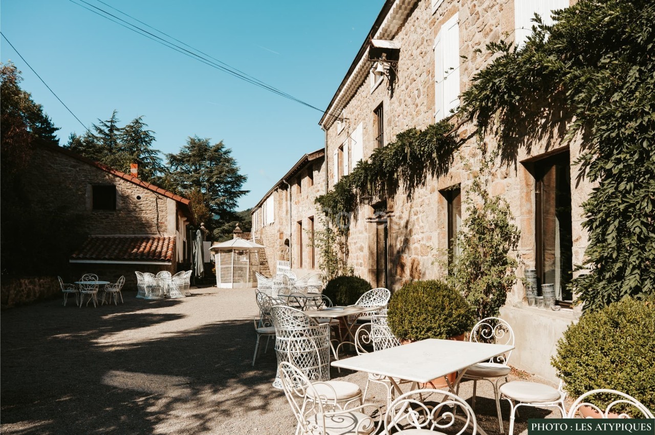 Cour extérieure ensoleillée avec tables et chaises en fer forgé devant un bâtiment en pierre couvert de végétation.