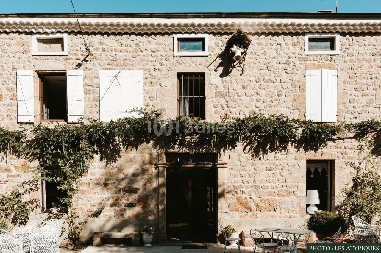 Façade en pierre d'une maison avec volets blancs, vigne grimpante et mobilier de jardin sur une terrasse.