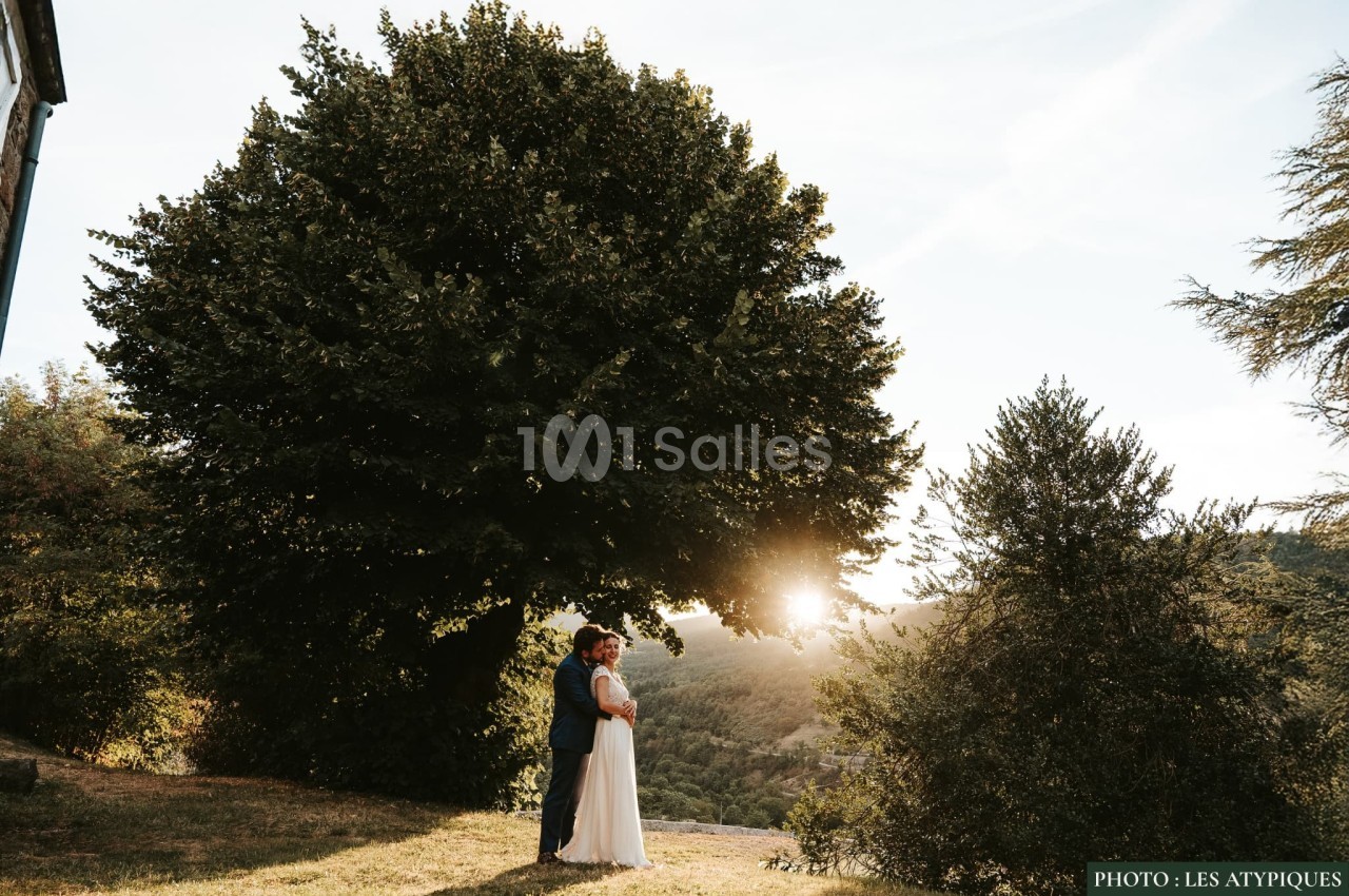 Un couple debout sous un grand arbre au coucher du soleil, entouré de verdure et de collines.