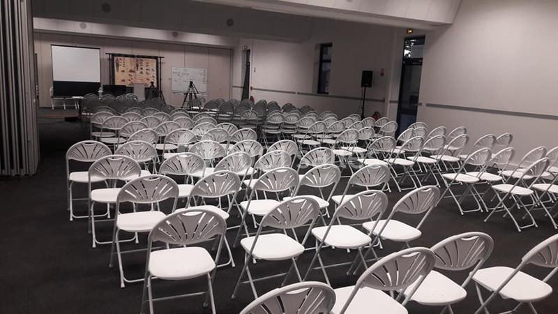 Salle de conférence vide avec des rangées de chaises blanches alignées face à un écran de projection.