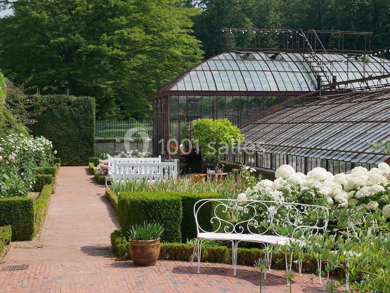 Serre en verre entourée de jardins fleuris, avec bancs blancs et allée en briques bordée de haies et de plantes.