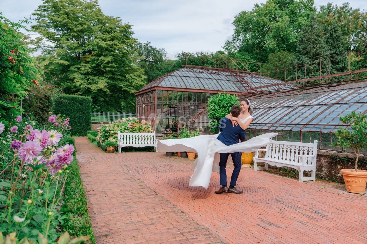 Un couple en tenue de mariage danse dans une allée pavée près de serres entourées de verdure et de fleurs.