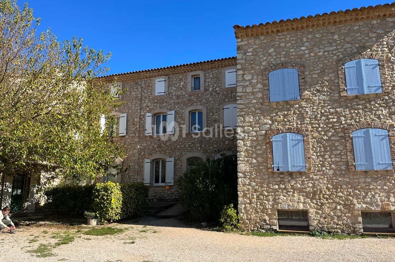 Bâtiment en pierre avec volets bleus, entouré d'arbres et donnant sur une cour ensoleillée.