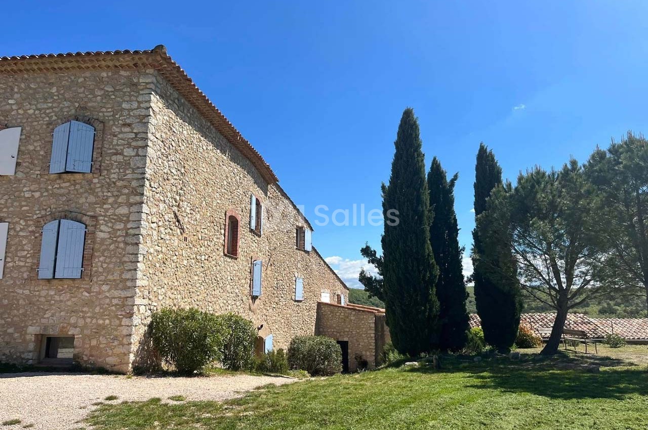 Bâtisse en pierre avec volets blancs, entourée de cyprès et d'arbres, sous un ciel bleu dégagé.