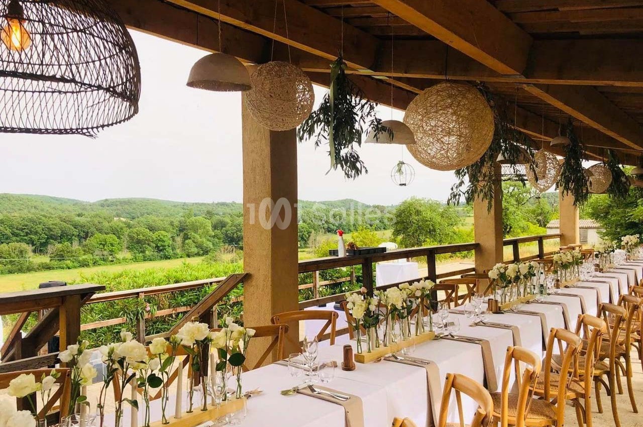 Tables décorées avec des fleurs blanches alignées sous une pergola en bois, offrant une vue sur un paysage verdoyant.