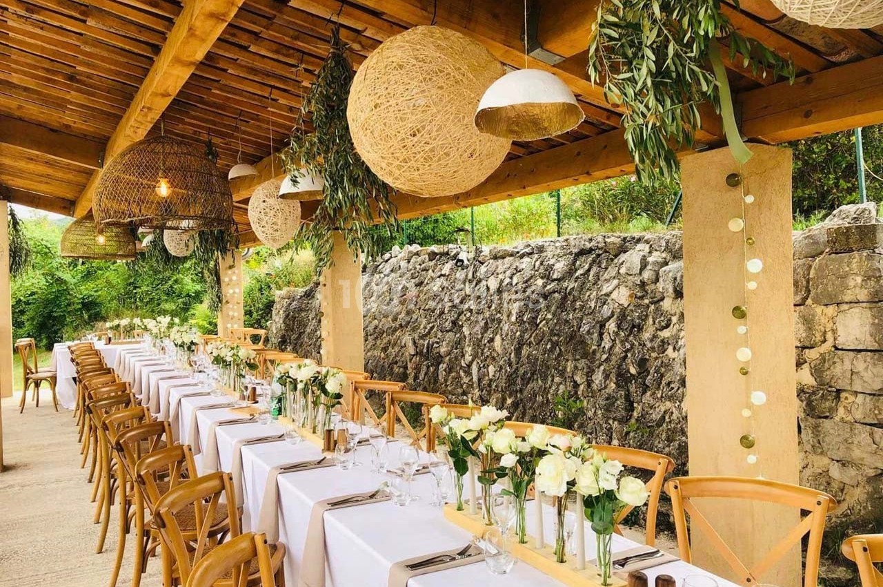 Tables longues décorées de nappes blanches, fleurs et chaises en bois sous une pergola ornée de lampes et feuillages.