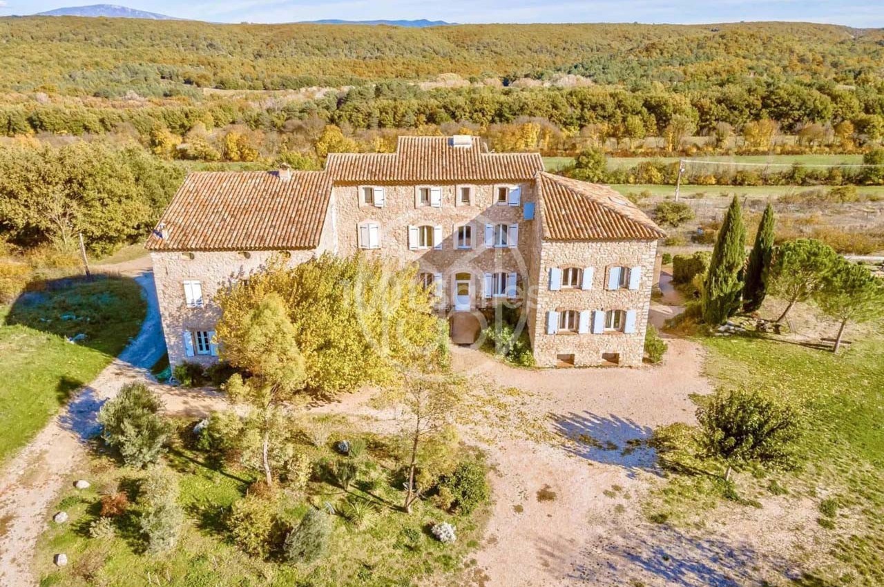 Vue aérienne d'une grande maison en pierre entourée de végétation, située dans un paysage rural.