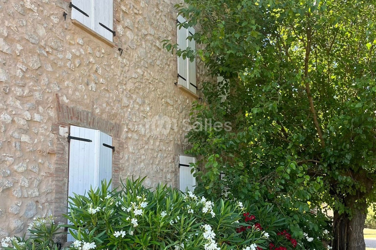 Façade en pierre d'une maison avec volets blancs, entourée de lauriers fleuris et d'un arbre verdoyant.
