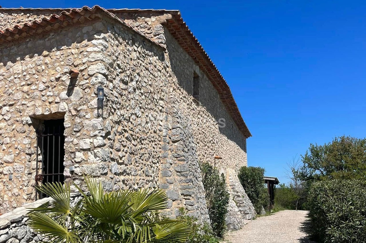 Bâtisse en pierre avec toit en tuiles, entourée de végétation et d'un chemin sous un ciel bleu clair.