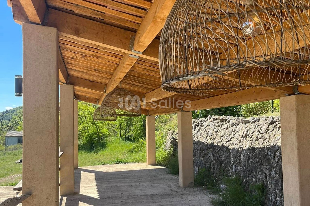 Structure en bois avec toit en tuiles, abritant des suspensions en osier, entourée de verdure et d'un muret en pierre.
