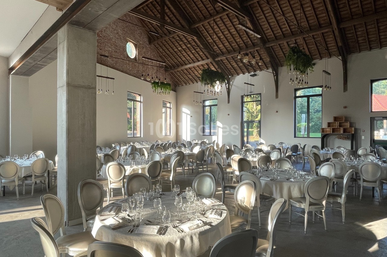 Salle de réception lumineuse avec tables rondes dressées, chaises élégantes et plafond en bois apparent.