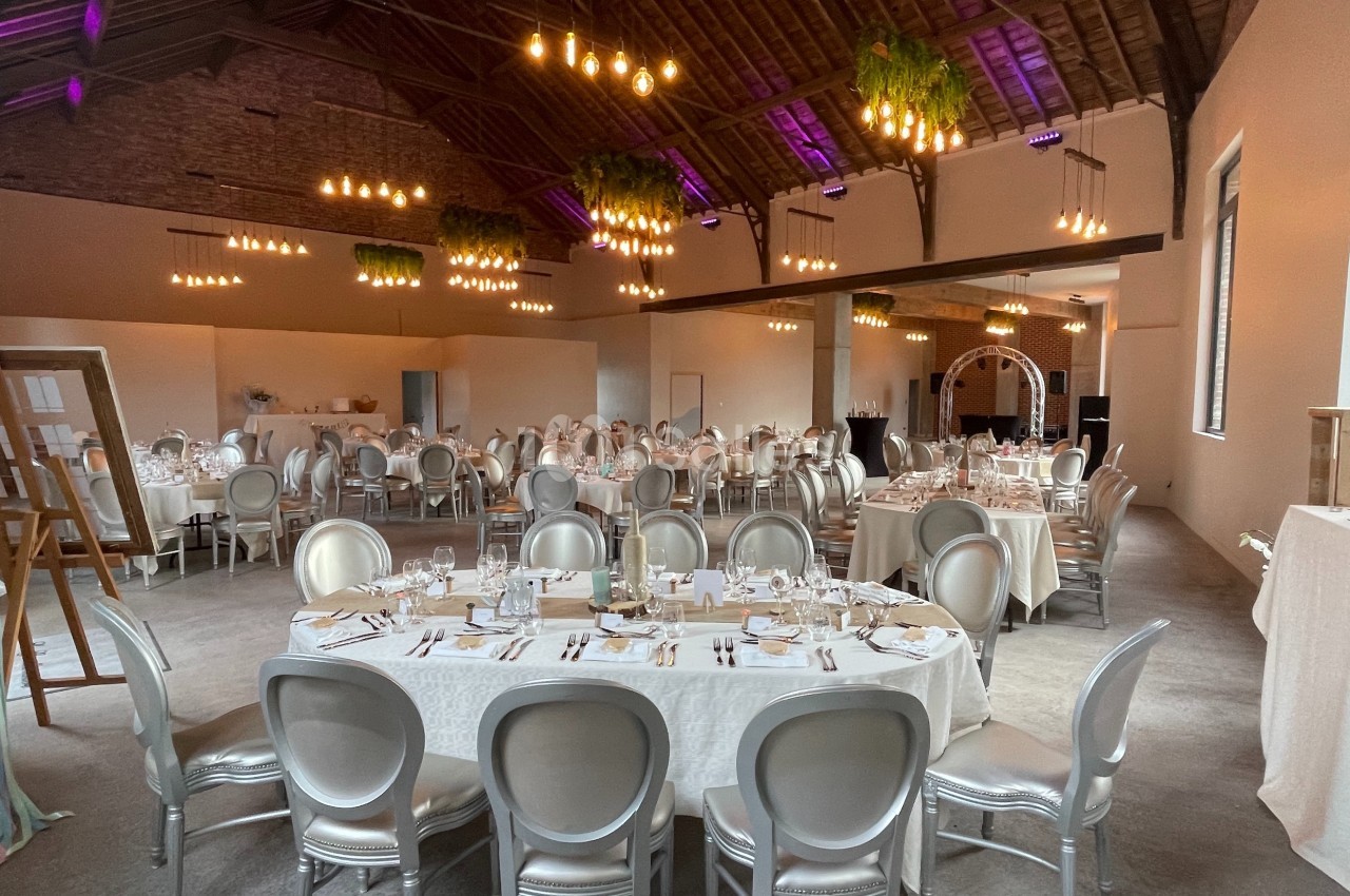 Salle de réception décorée avec des tables rondes dressées, des chaises argentées et des luminaires suspendus.