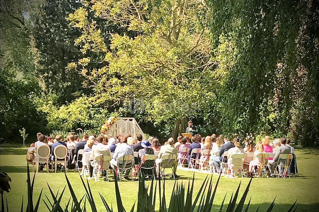 Cérémonie en plein air avec des invités assis devant un officiant, dans un jardin verdoyant et ensoleillé.