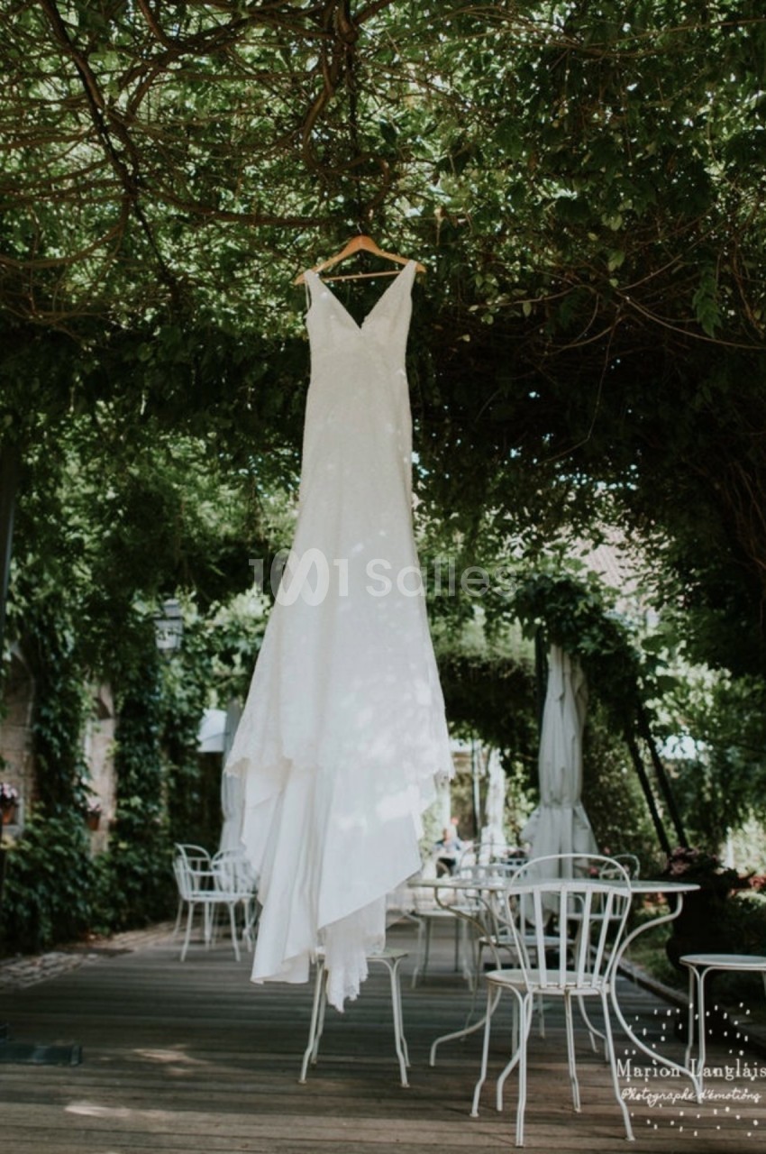 Robe de mariée blanche suspendue à une branche sous une pergola verdoyante avec des tables et chaises en fer forgé.
