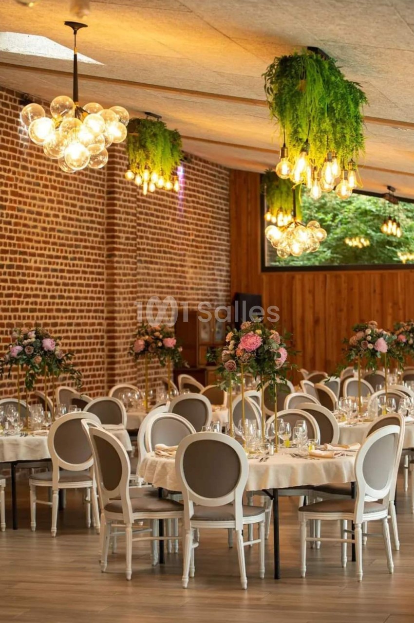 Salle de réception élégante avec murs en briques, tables rondes décorées de fleurs et luminaires suspendus.