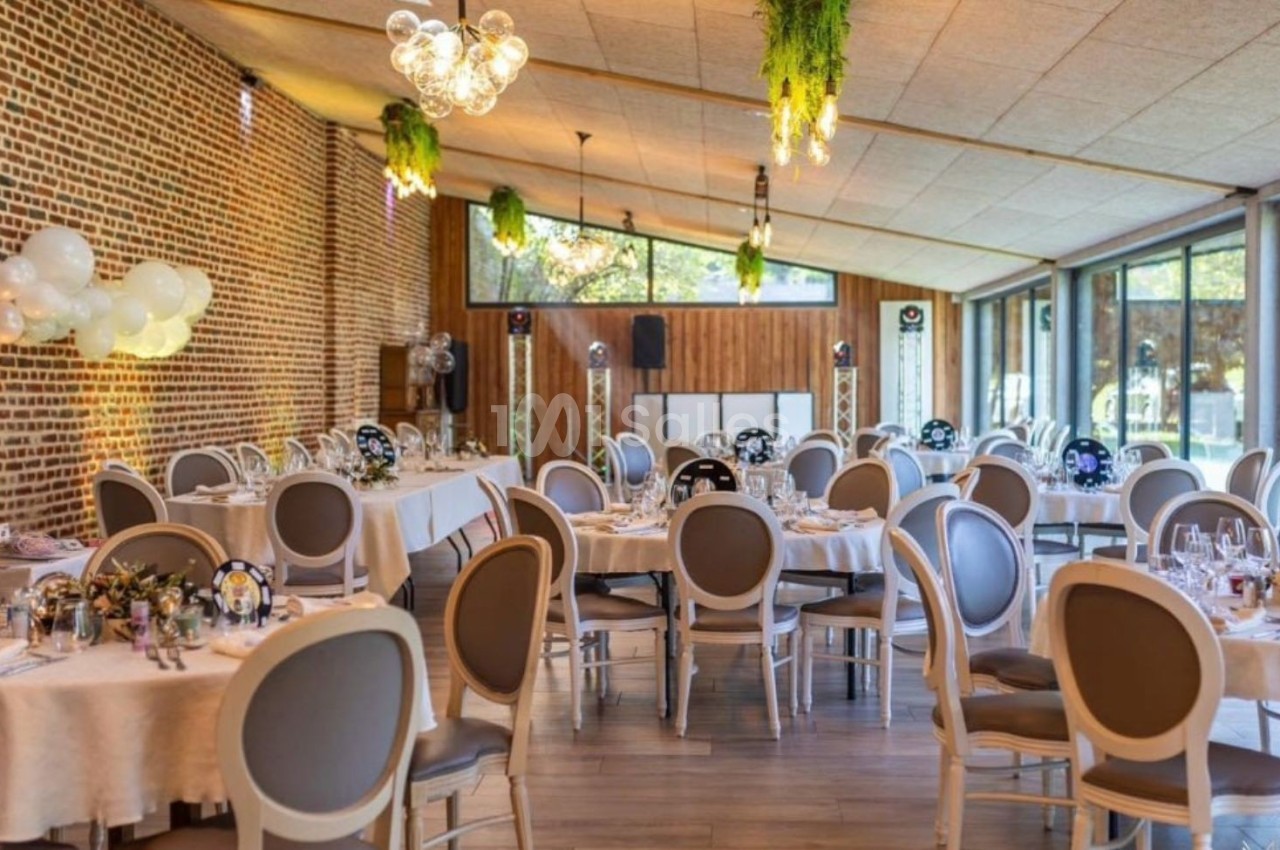 Salle de réception lumineuse avec tables rondes dressées, murs en briques, grandes baies vitrées et décorations suspendues.