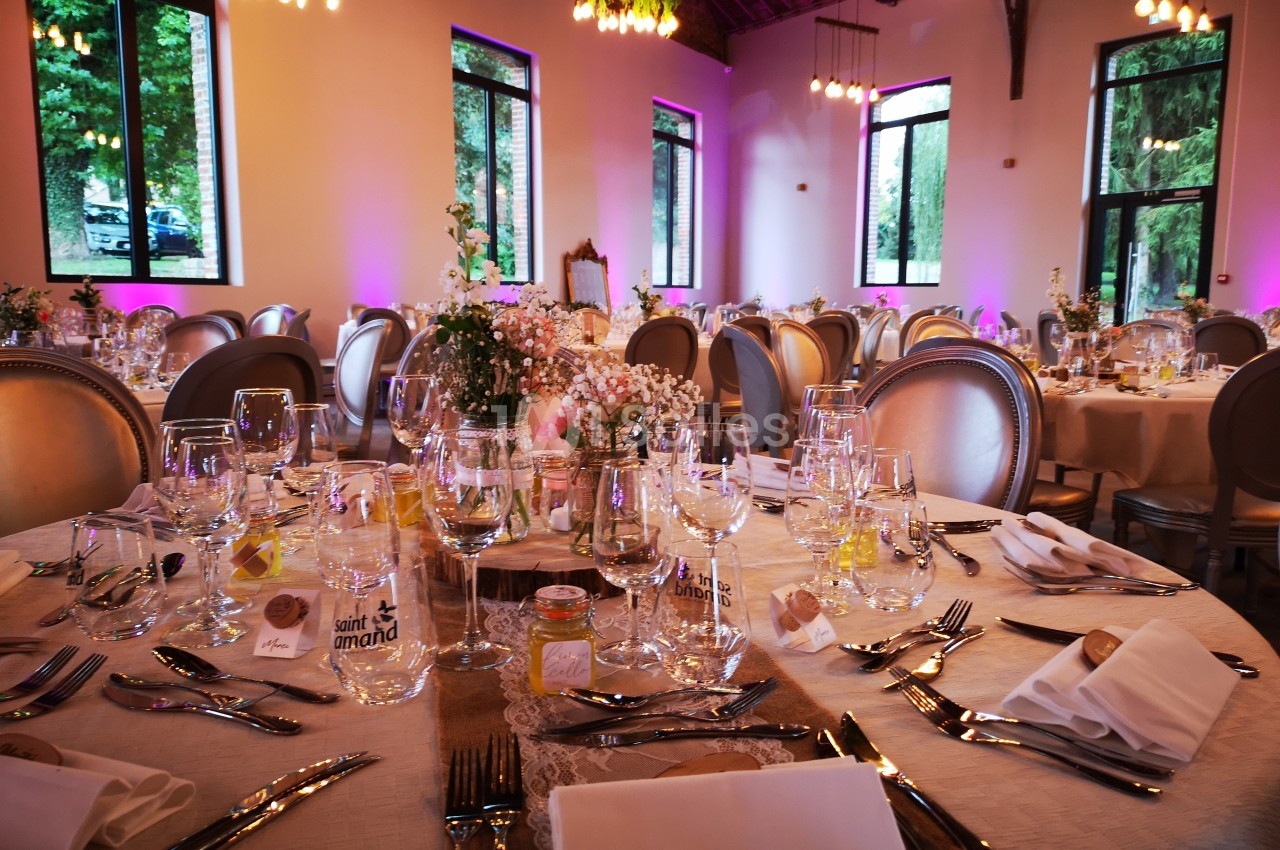 Salle de réception décorée avec des tables rondes élégamment dressées, éclairée par des lumières chaudes et des fenêtres…