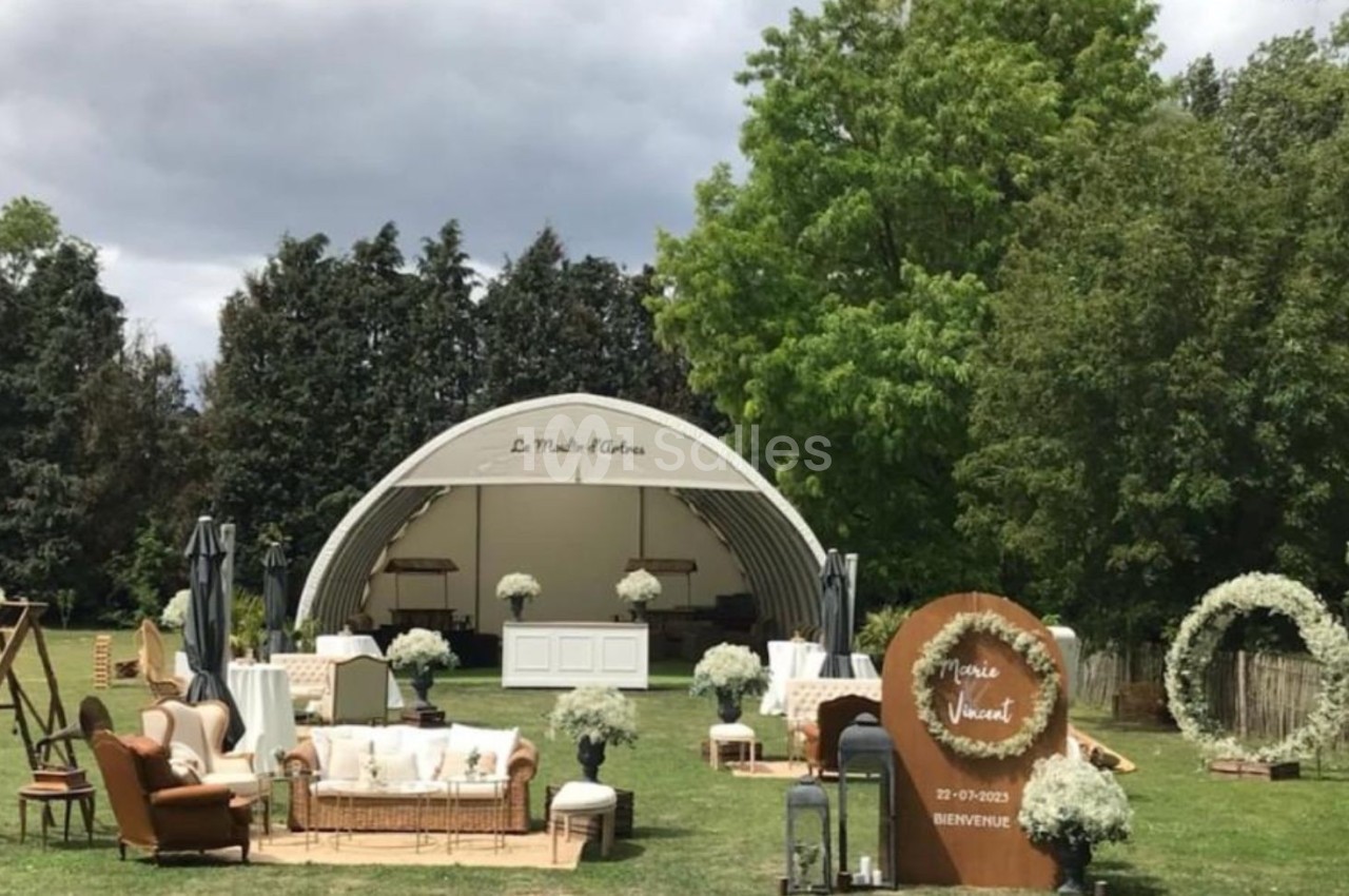 Espace extérieur aménagé pour un événement, avec une scène couverte, des fauteuils, des décorations florales et des arbres…
