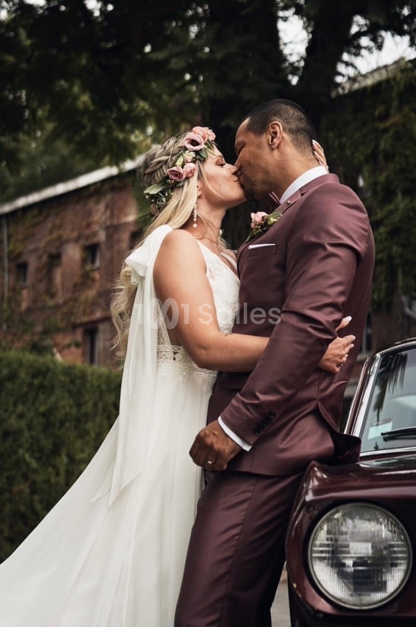 Un couple en tenue de mariage s'embrasse devant une voiture ancienne, avec un bâtiment en briques en arrière-plan.