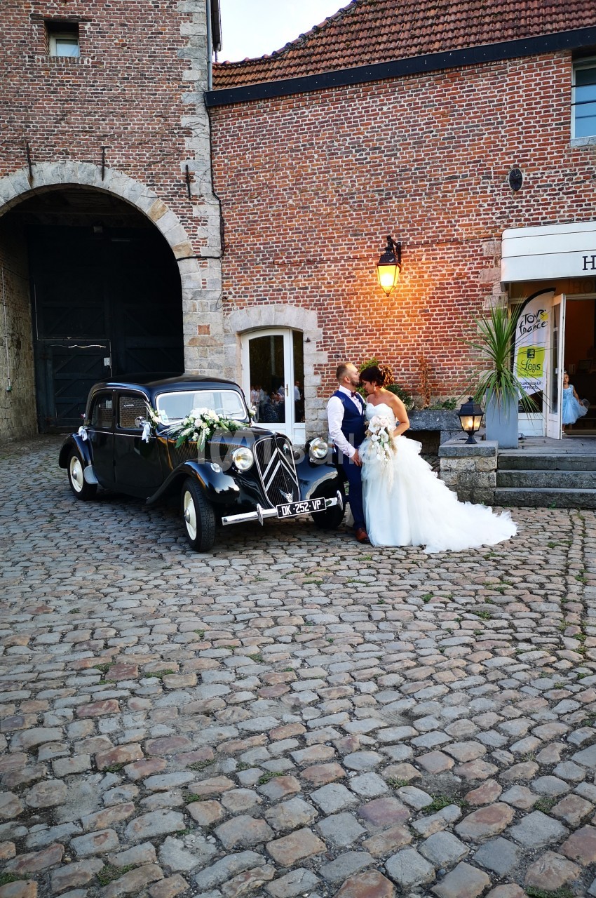 Un couple de mariés pose devant une voiture ancienne noire sur une cour pavée près d'un bâtiment en briques.