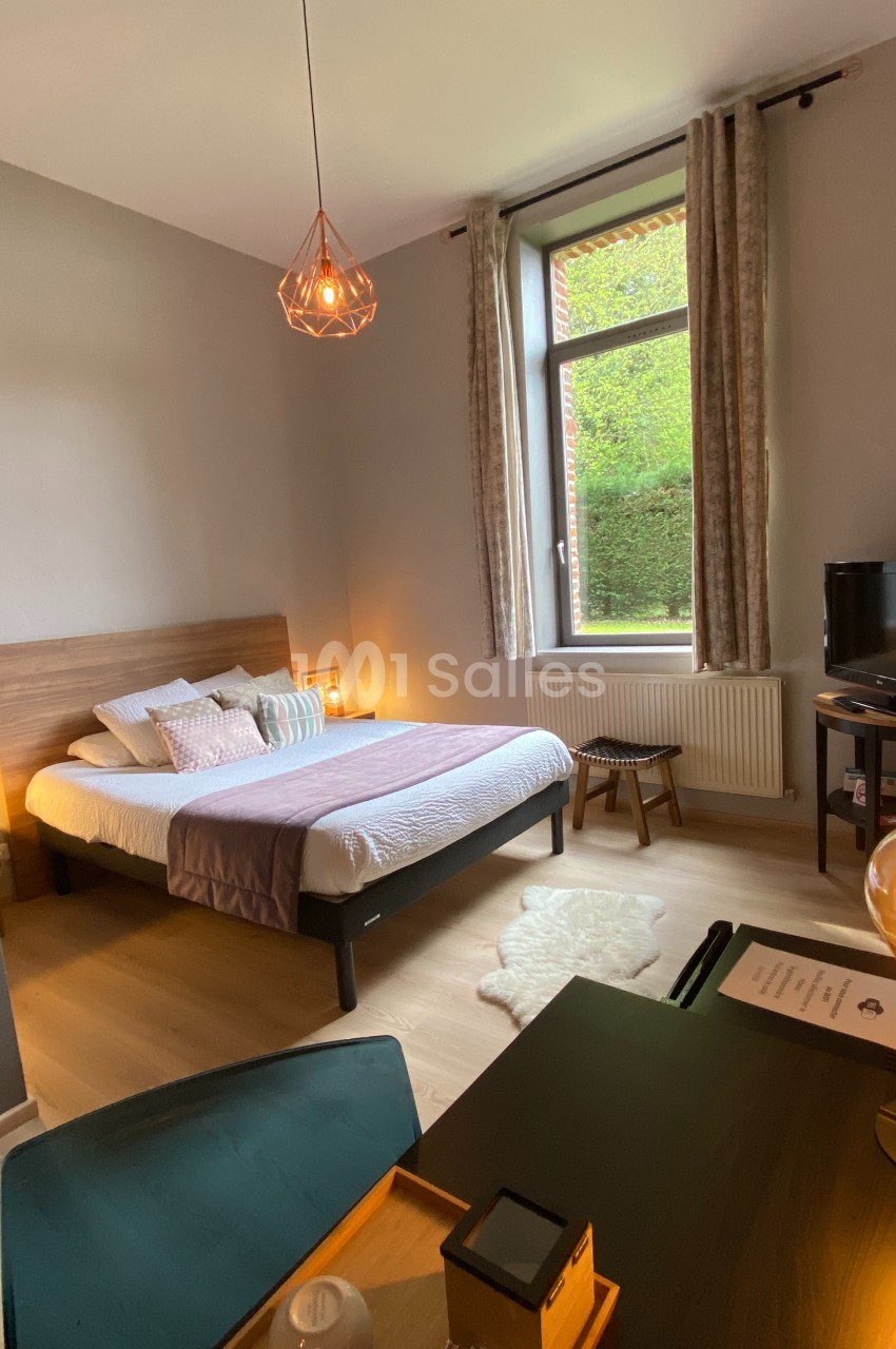 Chambre lumineuse avec lit double, bureau, lampe suspendue et grande fenêtre donnant sur un jardin verdoyant.