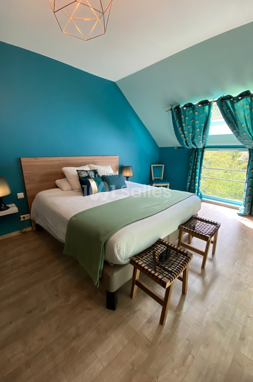 Chambre lumineuse avec lit double, murs bleus, rideaux assortis, parquet clair et vue sur un espace extérieur verdoyant.