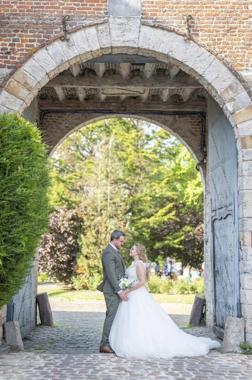 Un couple de mariés se tient sous une arche en pierre dans un jardin verdoyant par une journée ensoleillée.