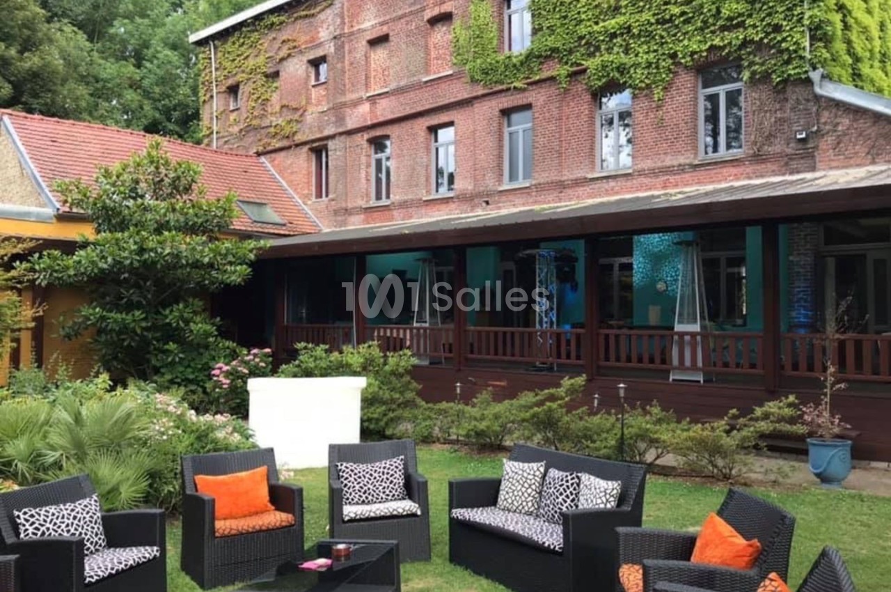 Façade en briques d'une maison avec terrasse couverte, jardin aménagé et mobilier de jardin en rotin noir.