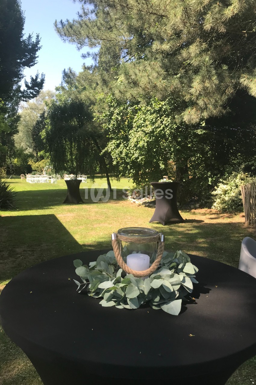 Table haute noire décorée de feuilles et d'une bougie dans un jardin verdoyant avec des tables en arrière-plan.