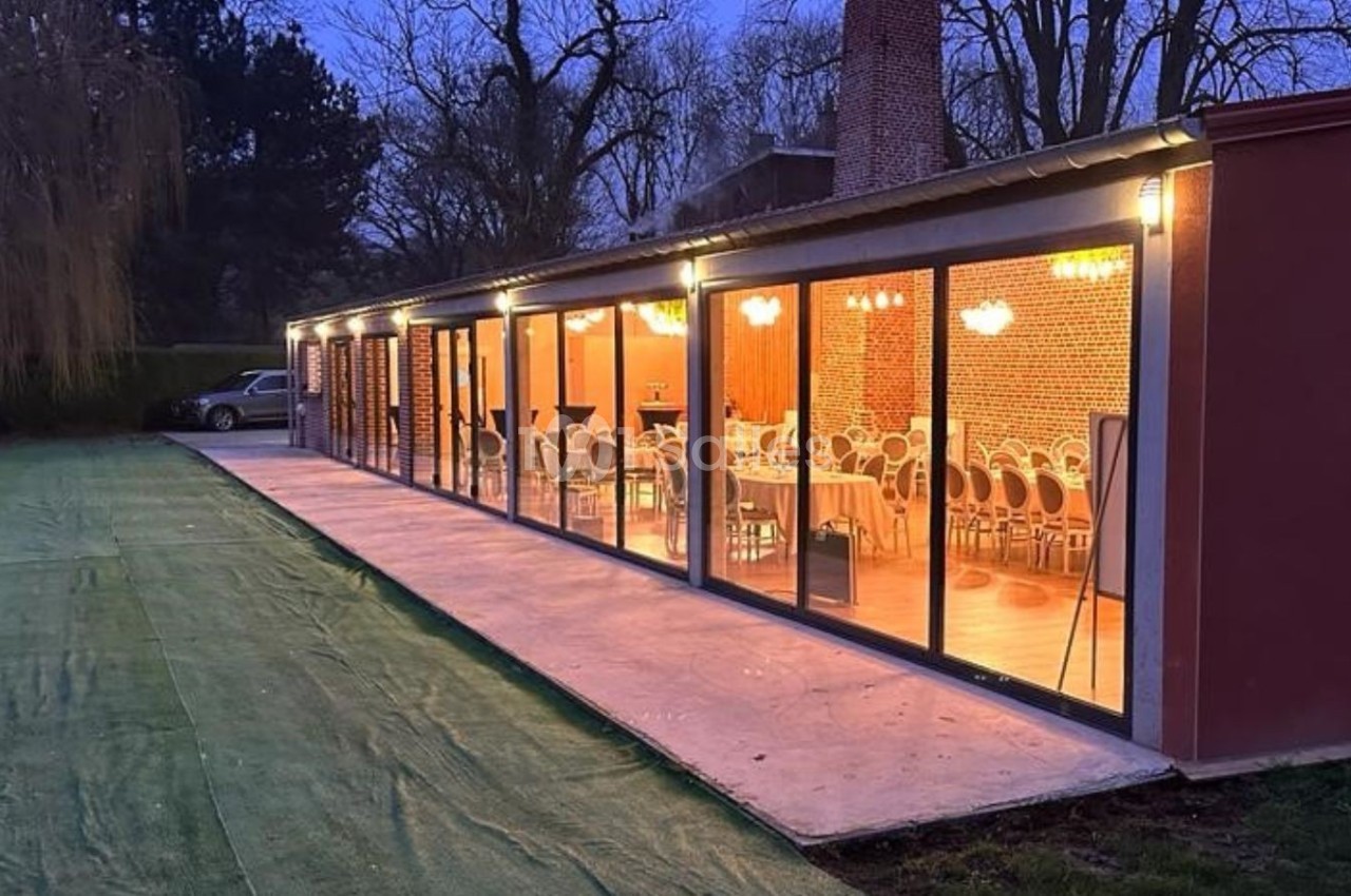 Salle vitrée éclairée en soirée, avec des tables et chaises visibles à l'intérieur, entourée d'un espace extérieur herbeux.