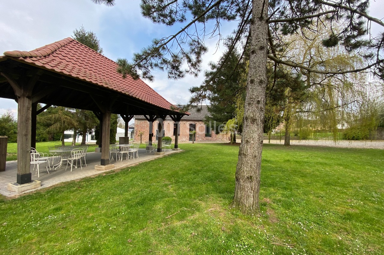Abri en bois avec toit en tuiles, entouré d'arbres et donnant sur une pelouse et un bâtiment en briques.