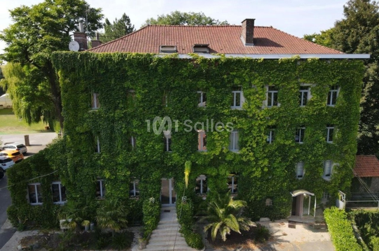 Façade d'un bâtiment recouverte de végétation dense, avec des fenêtres visibles et un toit en tuiles rouges.