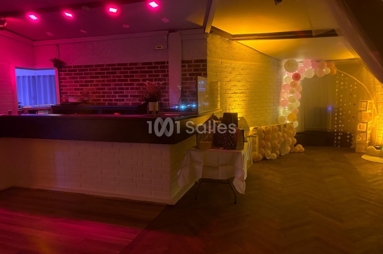 Salle décorée pour un événement, avec un bar, des lumières colorées et une arche de ballons blancs et dorés.