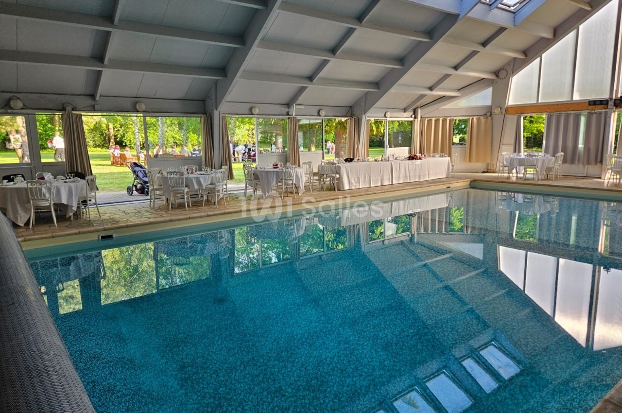 Piscine intérieure entourée de tables dressées pour un événement, avec vue sur un jardin à travers de grandes baies vitrées.