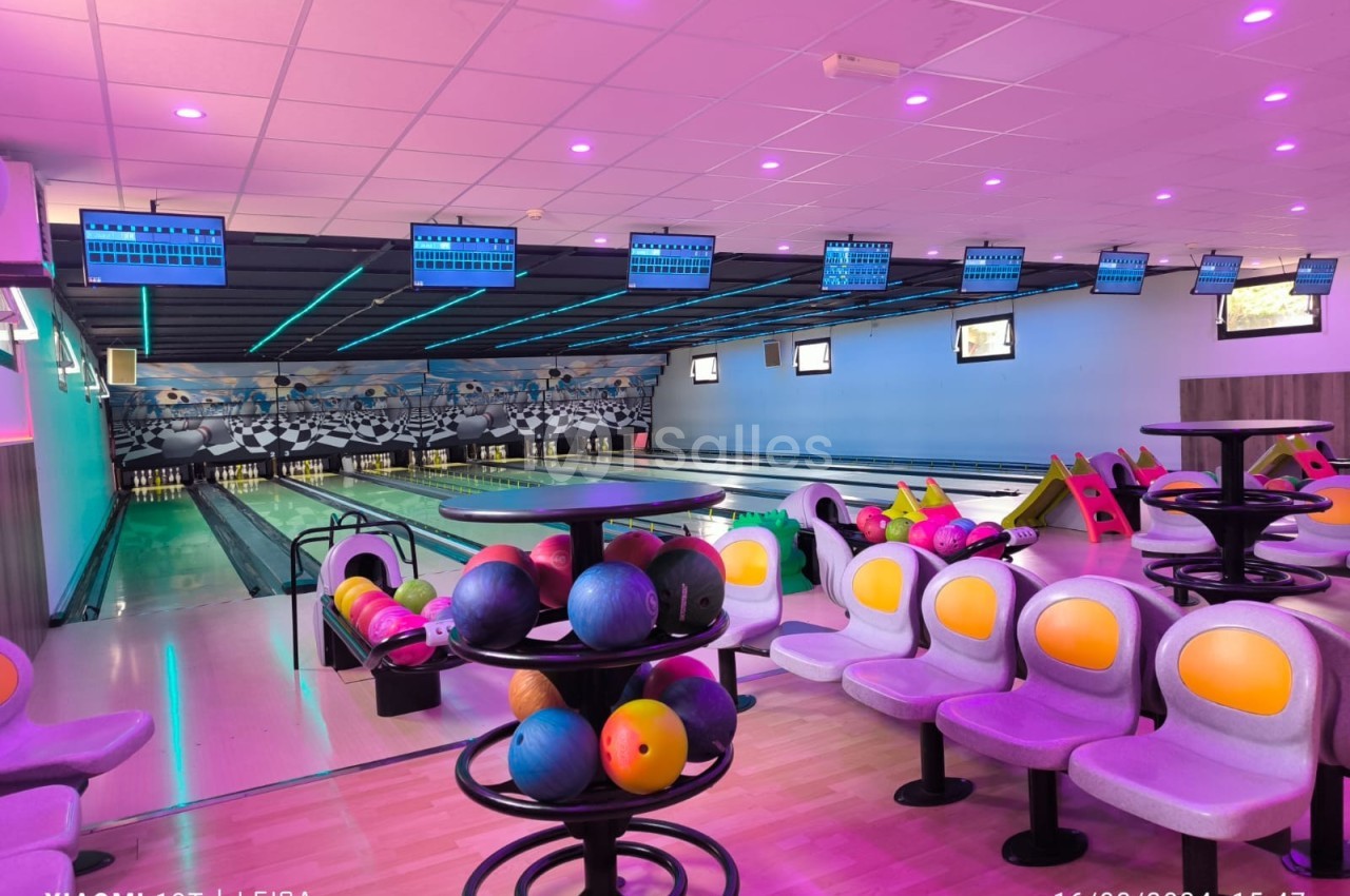 Salle de bowling avec plusieurs pistes, boules colorées sur des supports et sièges alignés sous un éclairage violet.