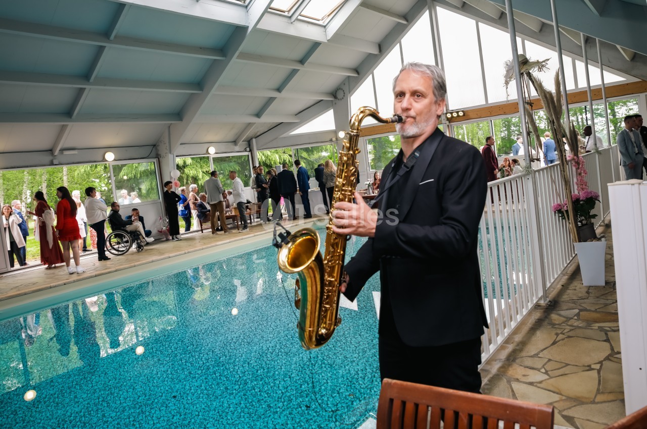 Un musicien joue du saxophone près d'une piscine intérieure lors d'un événement avec des invités en arrière-plan.