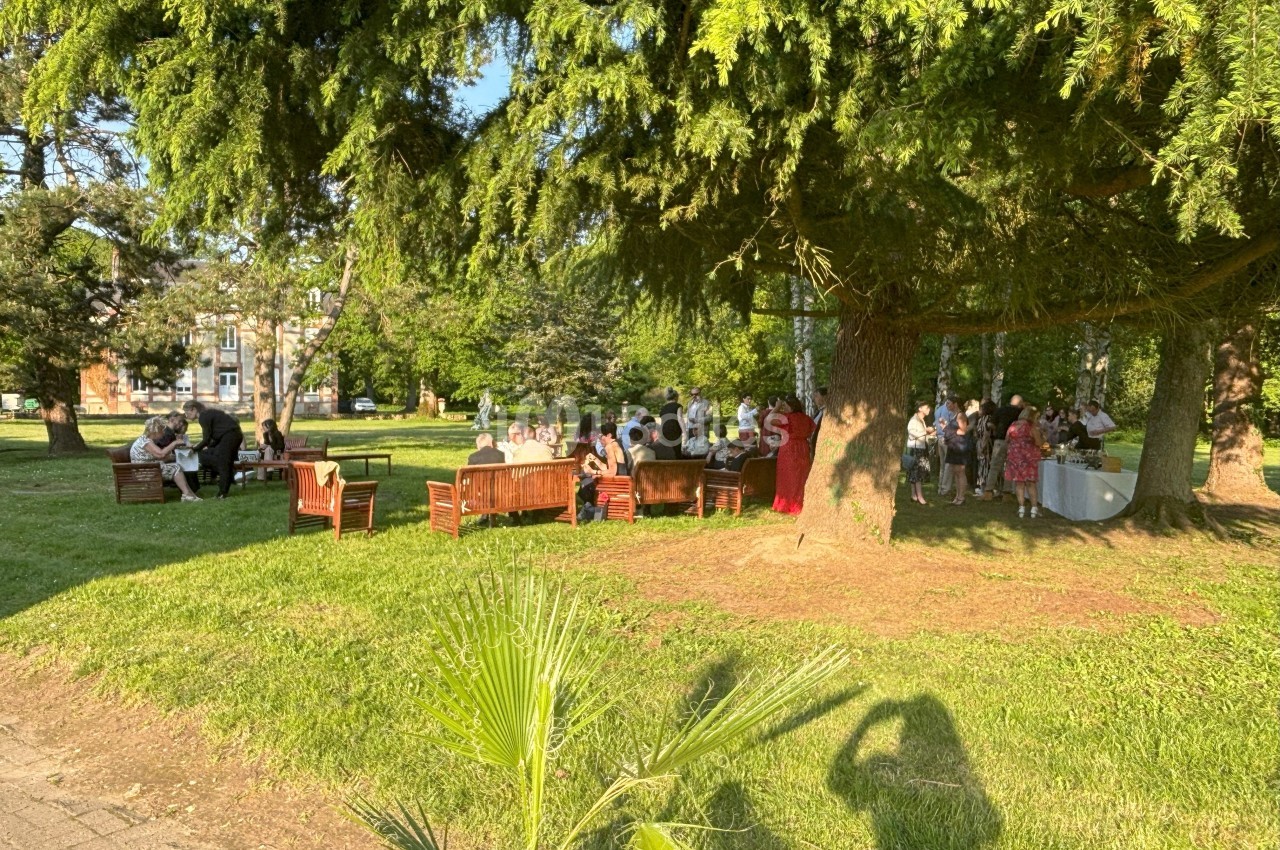 Groupe de personnes assises et debout dans un parc, près de tables et sous des arbres, par une journée ensoleillée.