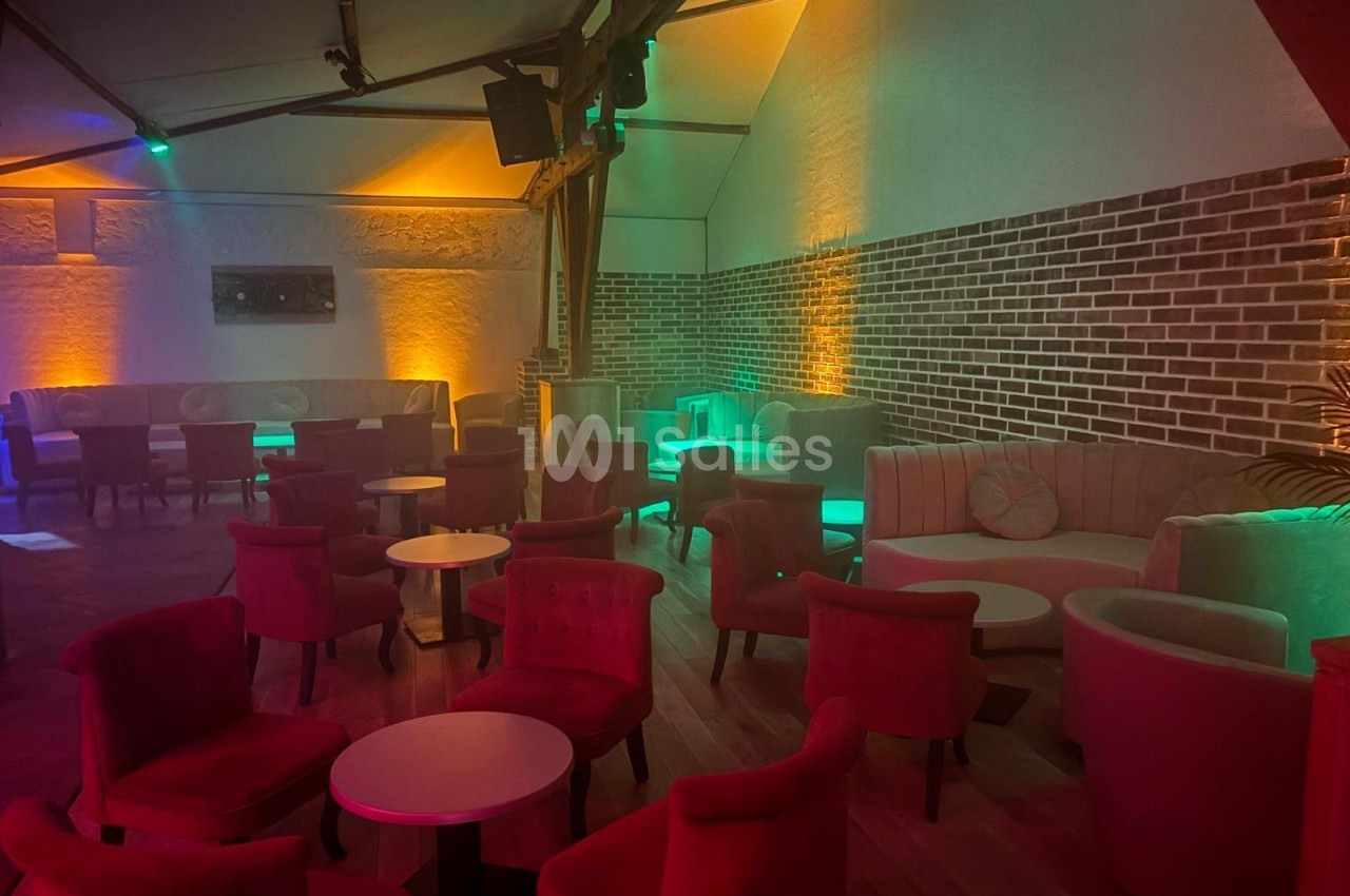 Salle avec des fauteuils rouges, tables basses et éclairage tamisé coloré, murs en briques et plafond incliné.