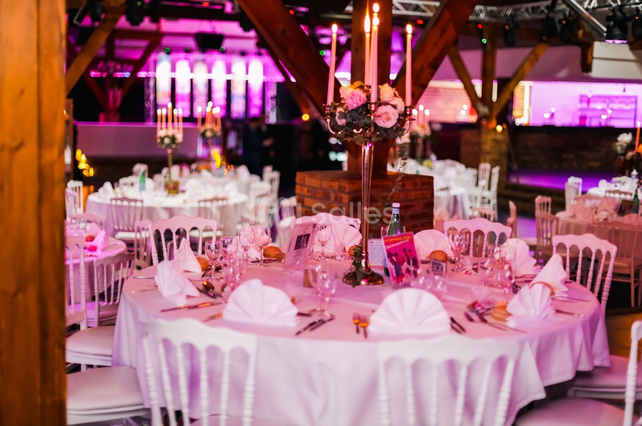 Salle de réception décorée avec des tables rondes dressées, nappes blanches, chandeliers et éclairage rose.